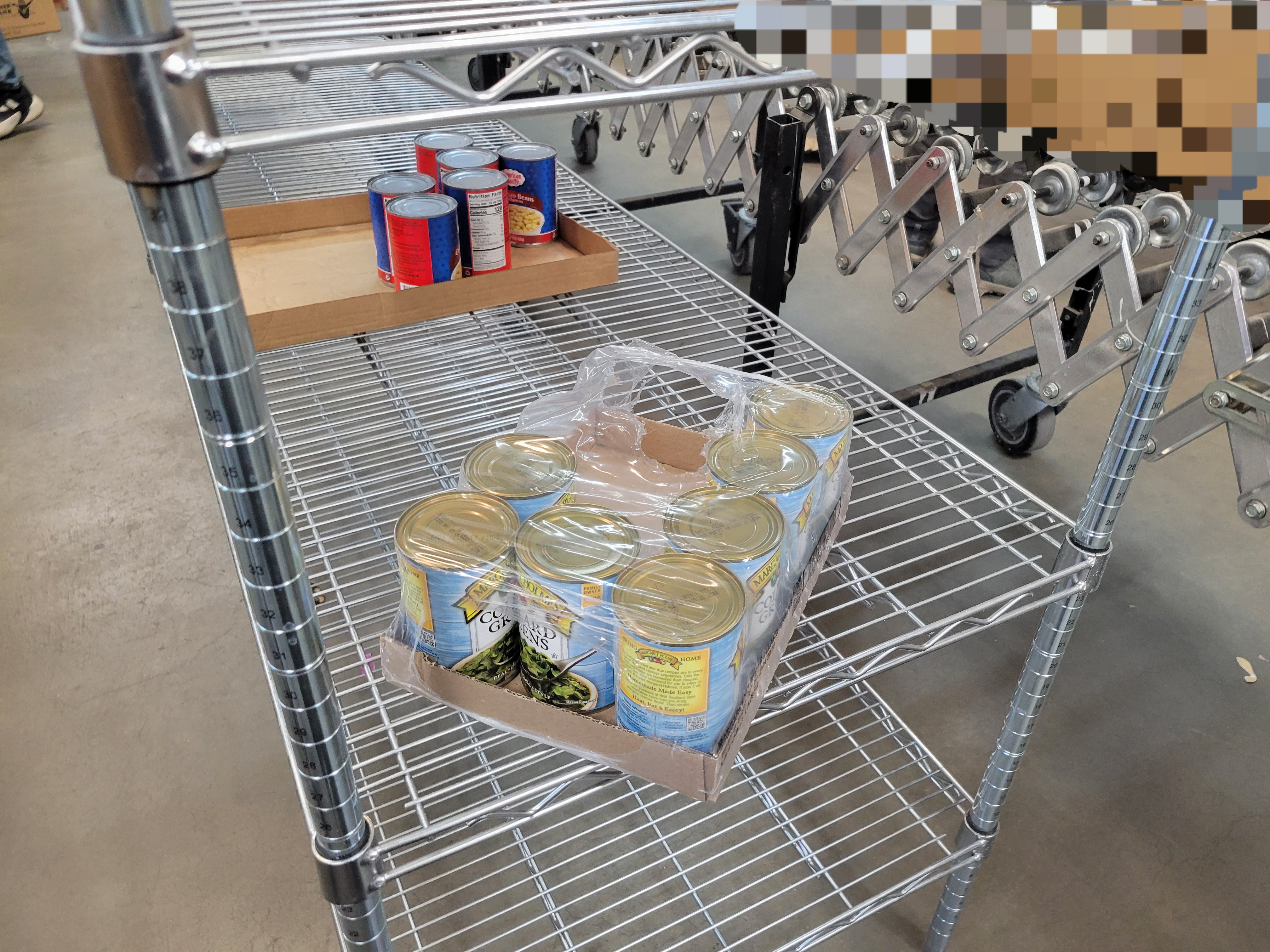 Cans of collard greens in plastic and box on a shelf and cans of black beans in cardboard near by.